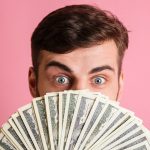 Close up portrait of a young man in white shirt covering his face with money banknotes and looking at camera isolated over pink background
